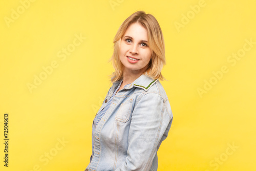 Profile portrait of pleased positive blonde woman wearing denim jacket being in good mood looking at camera with delighted face. Indoor studio shot isolated on yellow background.