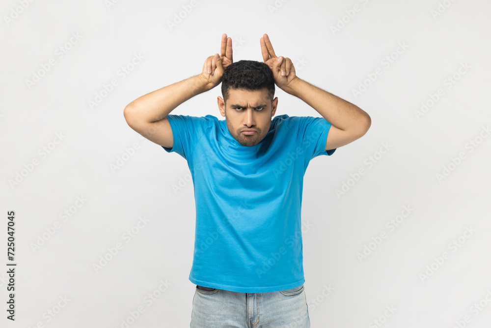 Portrait of angry strict handsome unshaven man wearing blue T- shirt standing showing bully gesture, looking at camera with aggression, attack. Indoor studio shot isolated on gray background. Portrait of angry strict handsome unshaven man wearing blue T- shirt standing showing bully gesture, looking at camera with aggression, attack. Indoor studio shot isolated on gray background.
