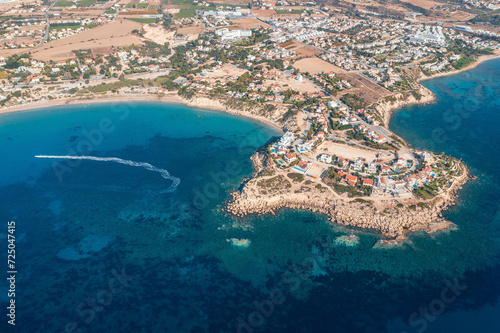 Aerial top down view of picturesque village nestled by Cyprus coastline, with azure waters and boats dotting seascape.