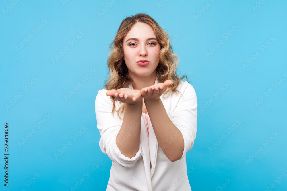 Portrait of flirting attractive blond woman with wavy hair sending air kissing over palm expressing romantic feelings, wearing white shirt. Indoor studio shot isolated on blue background. Portrait of flirting attractive blond woman with wavy hair sending air kissing over palm expressing romantic feelings, wearing white shirt. Indoor studio shot isolated on blue background.