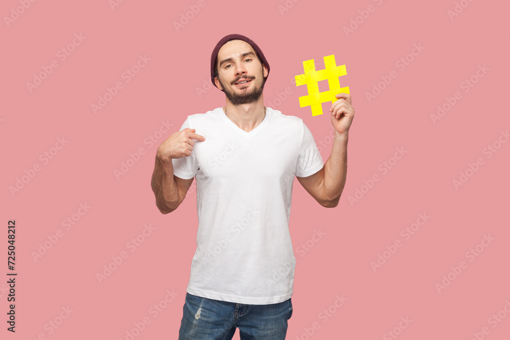 Portrait of smiling positive handsome bearded man in white T-shirt and beany hat standing, holding yellow hashtag and pointing at himself. Indoor studio shot isolated on pink background. Portrait of smiling positive handsome bearded man in white T-shirt and beany hat standing, holding yellow hashtag and pointing at himself. Indoor studio shot isolated on pink background.