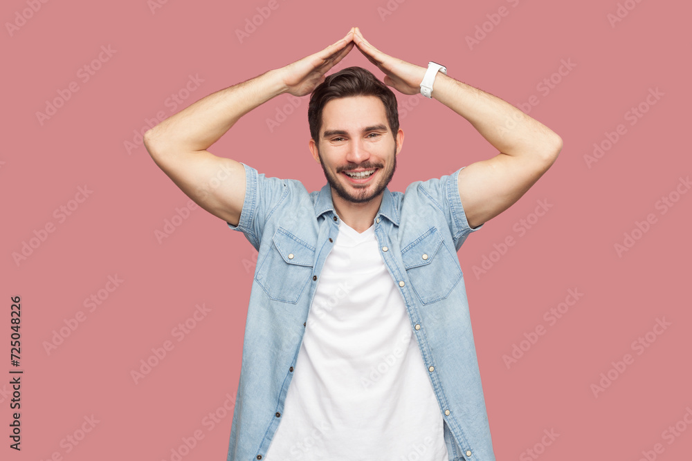 Satisfied happy bearded man in blue casual style shirt standing making house symbol, roof gesture over head and smiling, feeling safe protected. Indoor studio shot isolated on pink background. Satisfied happy bearded man in blue casual style shirt standing making house symbol, roof gesture over head and smiling, feeling safe protected. Indoor studio shot isolated on pink background.