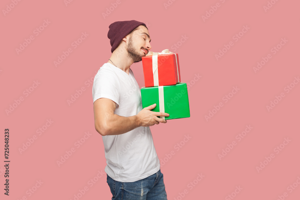 Portrait of satisfied bearded man in white T-shirt and beany hat standing with closed eyes, holding present boxes, celebrating holiday. Indoor studio shot isolated on pink background. Portrait of satisfied bearded man in white T-shirt and beany hat standing with closed eyes, holding present boxes, celebrating holiday. Indoor studio shot isolated on pink background.