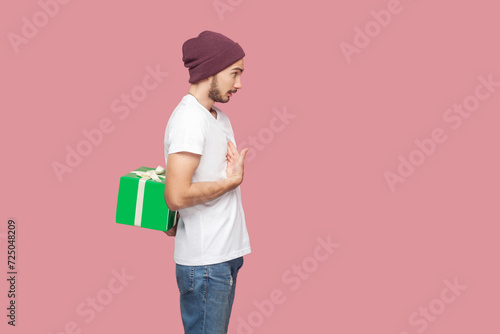 Side view portrait of surprised astonished bearded man in white T-shirt and beany hat standing pointing at himself, hiding present box behind. Indoor studio shot isolated on pink background. © khosrork