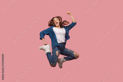Full length portrait of extremely happy brown haired woman jumping with clenched fists celebrating her successful achievements, wearing checkered shirt. Indoor studio shot isolated on pink background.