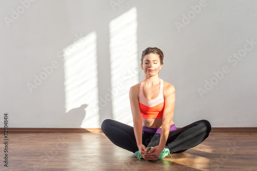 Portrait of attractive smiling woman sitting on floor stretching legs after hard workout, wearing black sports top and tights. Full length studio shot illuminated by sunlight from window.