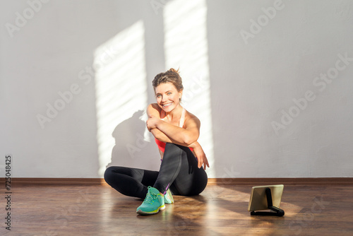Portrait of smiling confident woman siting resting after physical exercising, sport activity, wearing black sports top and tights. Full length studio shot illuminated by sunlight from window.