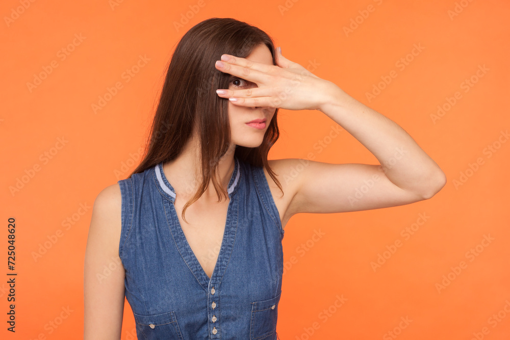 Portrait of scared shy brunette woman wearing denim dress peeking looking through fingers covering face sees something shameful. Indoor studio shot isolated on orange background. Portrait of scared shy brunette woman wearing denim dress peeking looking through fingers covering face sees something shameful. Indoor studio shot isolated on orange background.