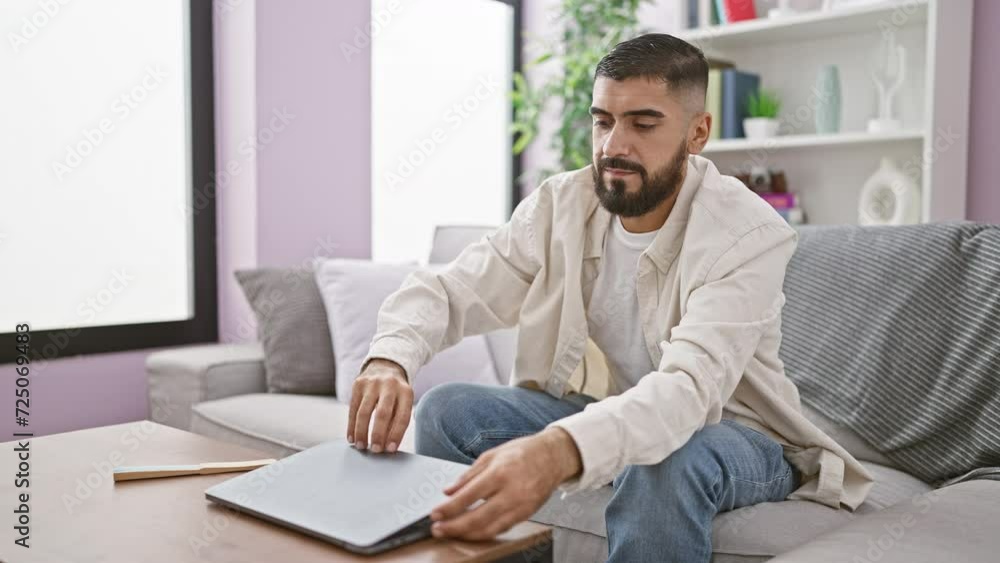 A bearded young man casually dressed using a laptop in a modern living room setting. A bearded young man casually dressed using a laptop in a modern living room setting.