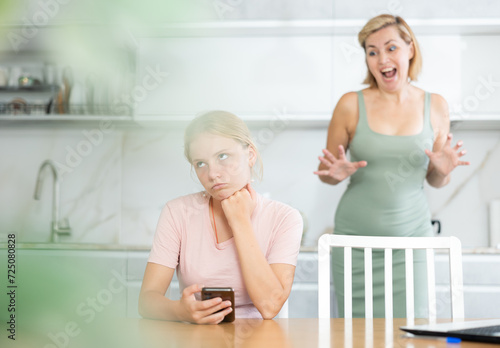 Bored girl is sitting in kitchen with phone in hands and listens to mothers claims, parents abuse for aimlessly spent time on social networks