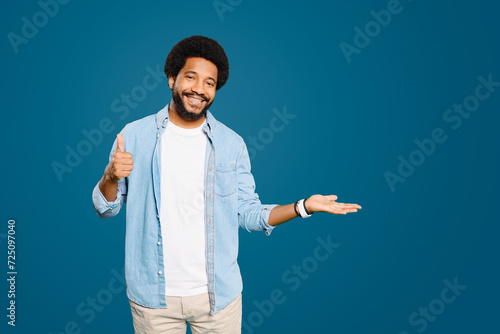Happy man gives a thumbs-up sign while maintaining an open palm with the other hand, suggesting approval and positivity and presenting something on a deep blue background. Copy-space, advertising