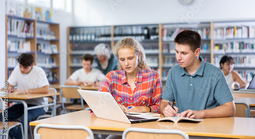 European fifteen-old-year schoolgirl, studying with a classmate in the school library on a laptop and making abstract in the copybook, preparing for lessons