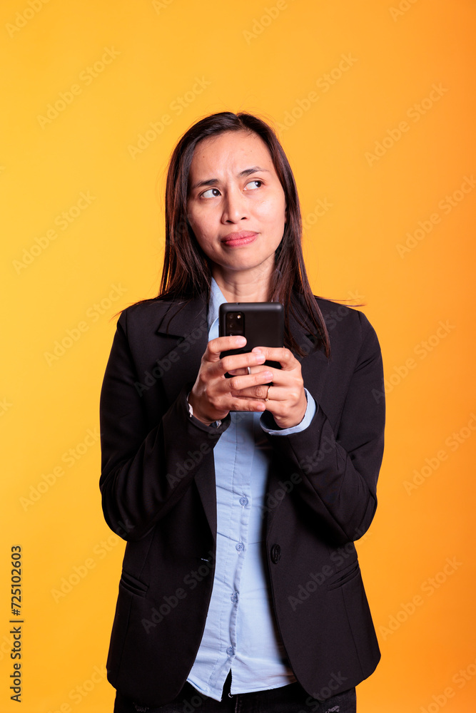 Pensive asian model typing message on smartphone, discussing with remote coworker in studio over yellow background. Cheerful woman in formal suit browsing information on internet, working at project Pensive asian model typing message on smartphone, discussing with remote coworker in studio over yellow background. Cheerful woman in formal suit browsing information on internet, working at project