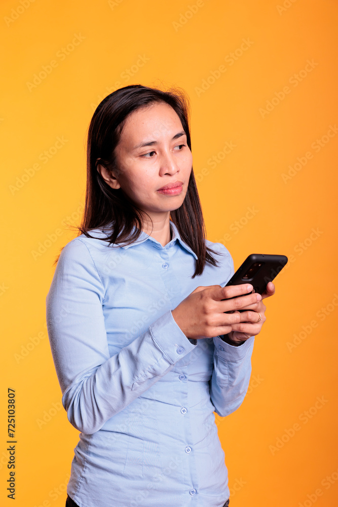 Pensive woman typing message on mobile phone, discussing with remote friend in studio over yellow background. Cheerful filipino young adult browsing on internet, searching information for project Pensive woman typing message on mobile phone, discussing with remote friend in studio over yellow background. Cheerful filipino young adult browsing on internet, searching information for project