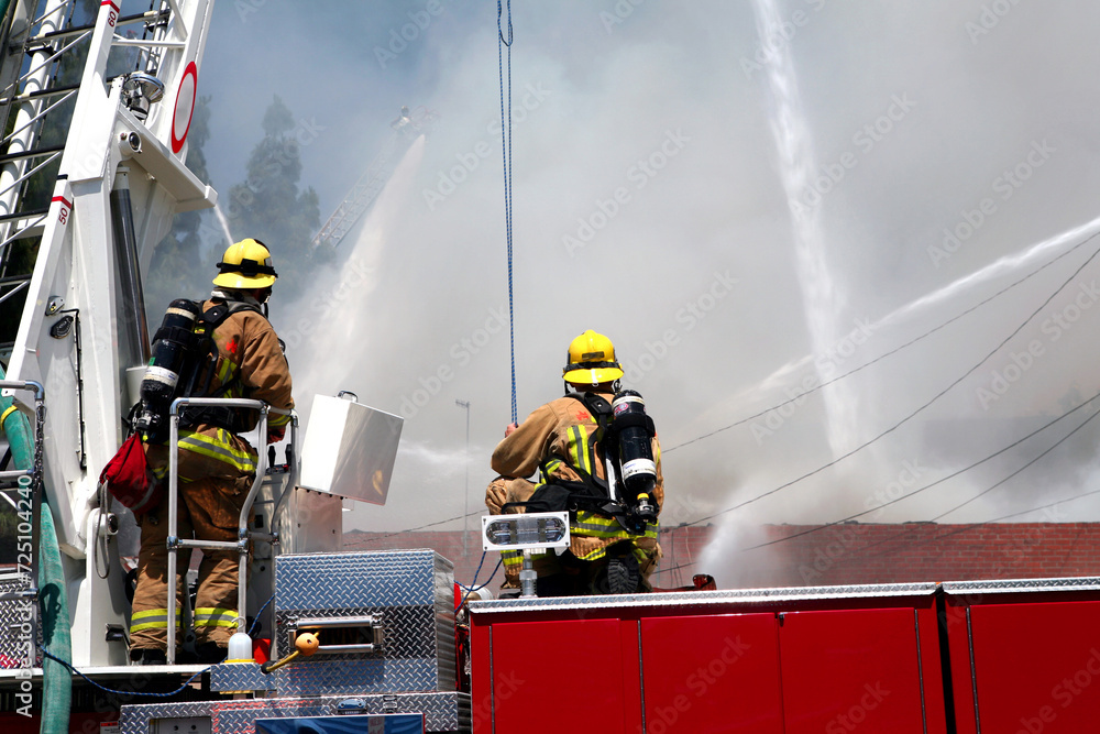 Fototapeta premium Firemen standing on a hook and ladder fire engine watching as water canons blast a structure fire with streams of water.