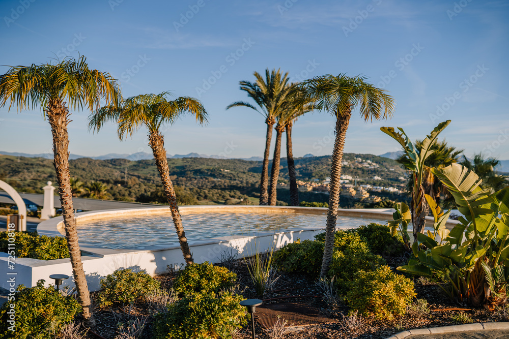 Sotogrante, Spain - January 25, 2024 - Palm trees and plants in the foreground with a reflective pool and hilly landscape in the background.