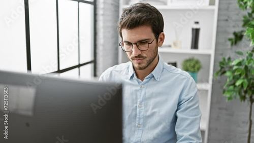 Handsome young hispanic man with beard feels frustrated in modern office setting, taking off glasses. photo
