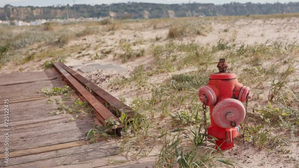 Rusty red fire hydrant on a sandy beach with wooden pathway and grass. Concept of safety, preparation, and contrast between man-made and natural environments. Rusty red fire hydrant on a sandy beach with wooden pathway and grass. Concept of safety, preparation, and contrast between man-made and natural environments.
