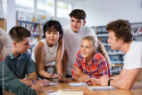 Group of fifteen-year-old schoolchildren are preparing for classes in the school library, discussing something and making notes in copybooks