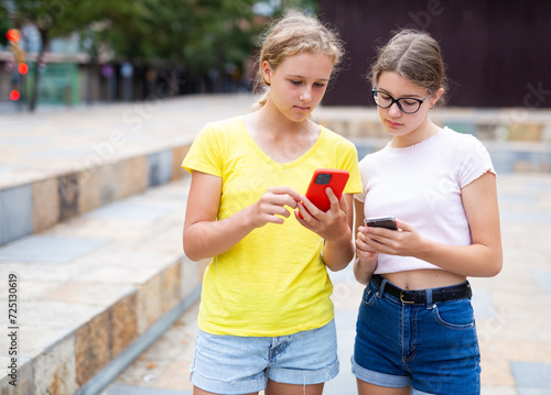 Two young girls standing in park and using their smartphones.
