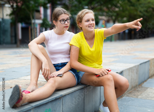 Two friendly girls sitting in square. Girl showing something to her friend by pointing index finger.