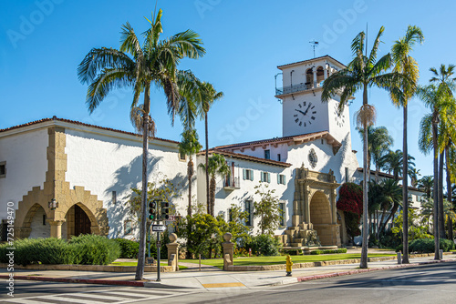 Santa Barbara, CA, USA - November 30, 2023: Santa Barbara County Courthouse, white stone and tower with palm trees and green lawn seen from SW corner against blue sky. Traffic light photo
