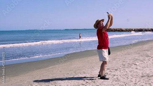 Older man wearing a vueltiao hat, red T-shirt and shorts, taking a selfie on the beautiful beaches of Cartagena de Indias, Colombia. photo