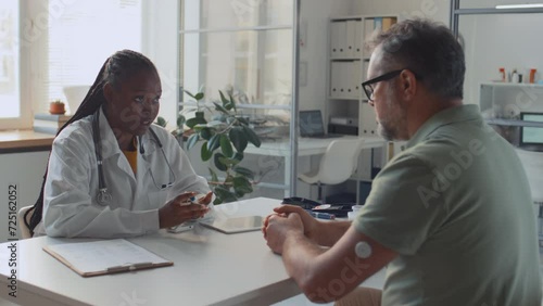 Medium shot of African American female medical doctor sitting at desk in clinic, consulting middle-aged Caucasian patient with diabetes, and demonstrating blood glucose control method photo