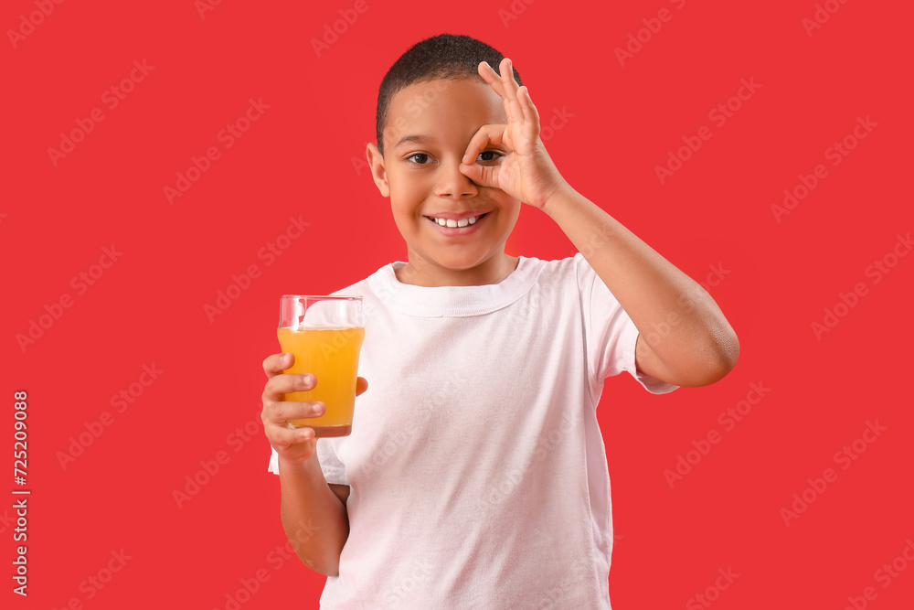 Little African-American boy with glass of orange juice showing OK on red background Little African-American boy with glass of orange juice showing OK on red background