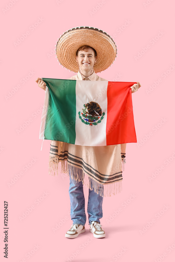 Young man in sombrero and poncho holding Mexican flag on pink background