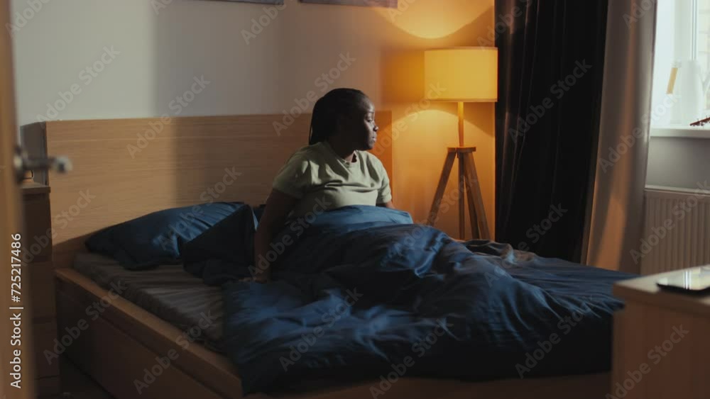 Full shot of young African American woman asleep alone in double bed at home in morning, then waking, sitting up, stretching and touching blood glucose monitoring device on upper arm Full shot of young African American woman asleep alone in double bed at home in morning, then waking, sitting up, stretching and touching blood glucose monitoring device on upper arm