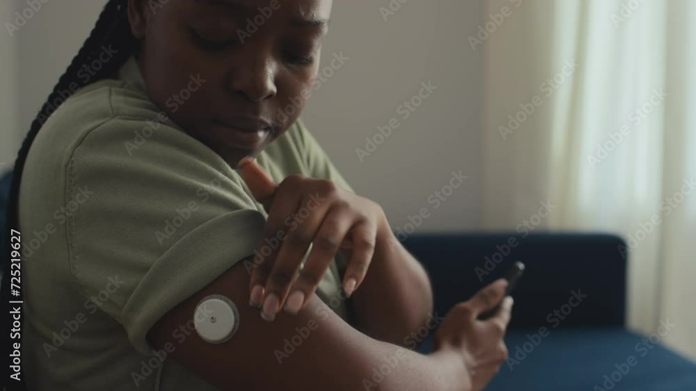 Medium close-up of young African American woman with diabetes sitting on couch in living room, touching blood sugar monitoring device on arm, then checking data on smartphone app Medium close-up of young African American woman with diabetes sitting on couch in living room, touching blood sugar monitoring device on arm, then checking data on smartphone app