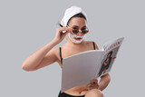 Beautiful young woman in sunglasses with shaving foam on her face and razor reading magazine on grey background