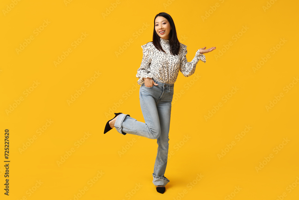 Beautiful young happy Asian woman in stylish jeans pointing at something on yellow background Beautiful young happy Asian woman in stylish jeans pointing at something on yellow background