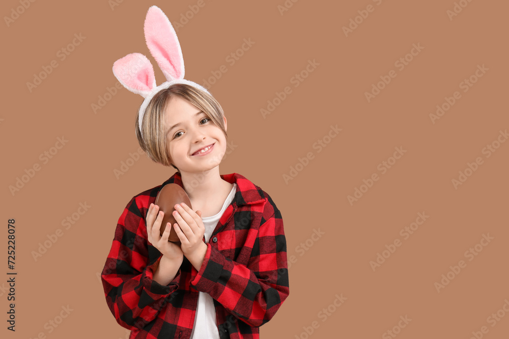 Happy little boy in bunny ears with chocolate egg on brown background. Easter celebration