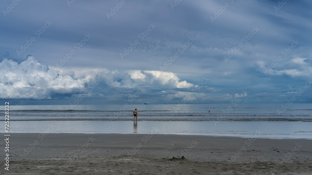 The waves of the turquoise ocean roll towards the shore, foaming and spreading over the sandy beach. A lonely man goes into the sea. Reflection. A ship is on the horizon. Clouds in the blue sky.  