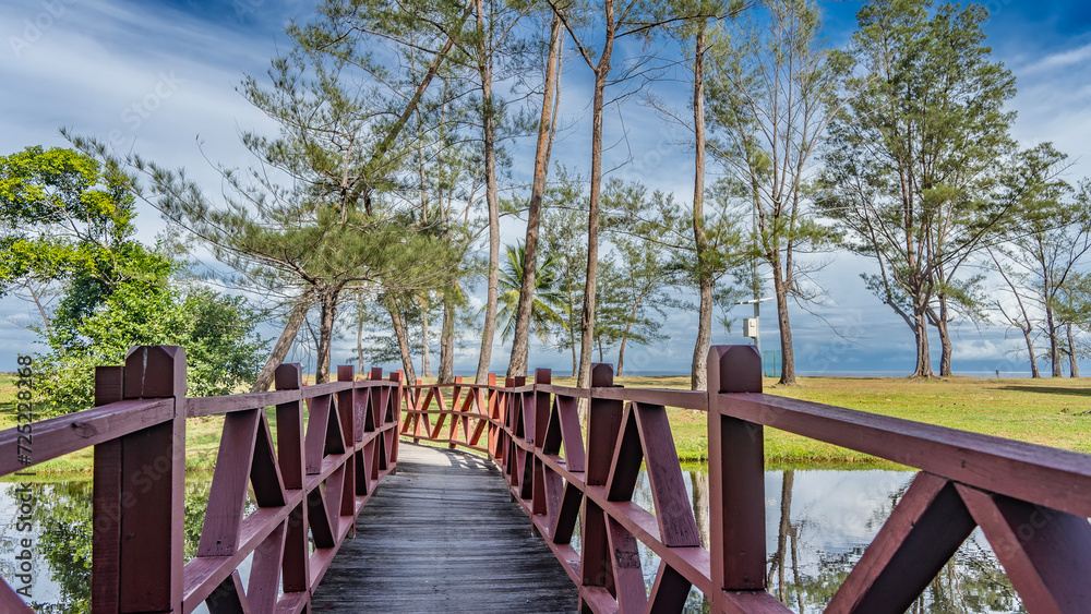 A plank pedestrian bridge with wooden railings runs over a calm river. The ocean is ahead. Green trees against a blue sky and clouds. Reflection in the water. Malaysia. Borneo. Kota Kinabalu