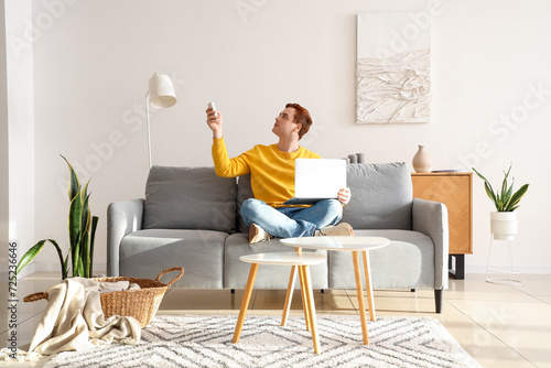 Young man with laptop turning on air conditioner on couch in living room photo