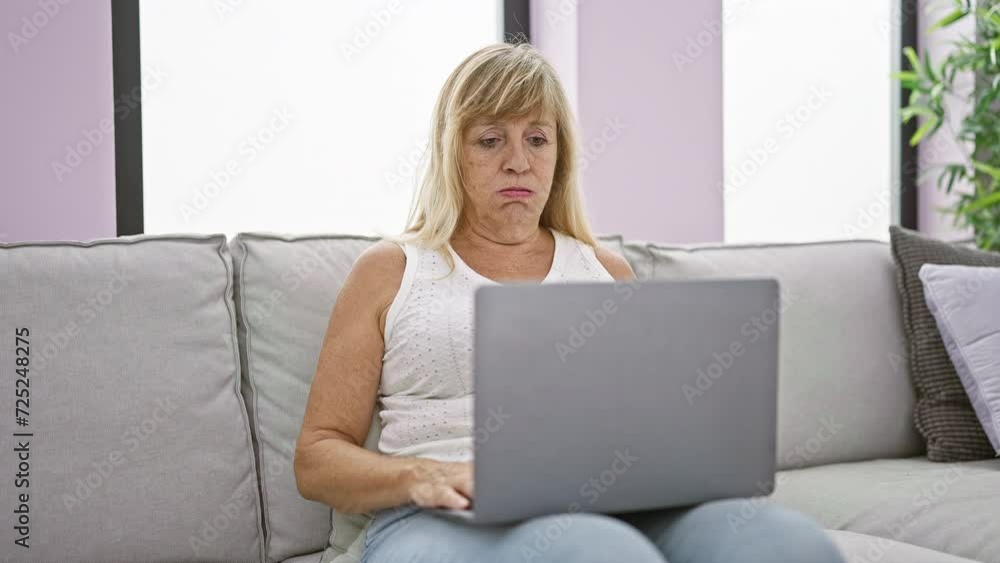 In her cozy home, a serious and focused middle age blonde woman is sitting on the sofa, totally absorbed, using her laptop in the living room's welcoming interior. In her cozy home, a serious and focused middle age blonde woman is sitting on the sofa, totally absorbed, using her laptop in the living room's welcoming interior.