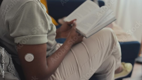 Medium side shot of midsection of anonymous black female with diabetes reading book on couch at home, touching glucose monitoring sensor on arm photo