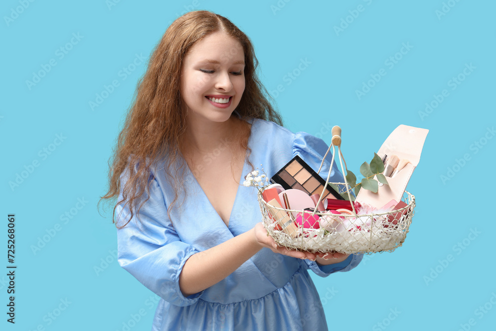 Beautiful young happy woman with basket full of decorative cosmetics on blue background