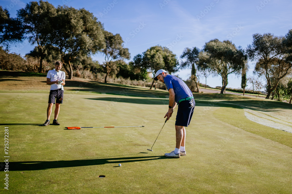 Obraz premium Sotogrante, Spain - January 25, 2024 - Two men on a golf green, one putting towards the hole, the other observing, with trees in the background.