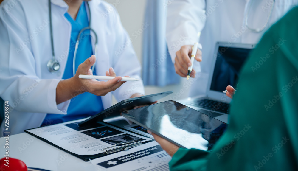 Medical team having a meeting with doctors in white lab coats and surgical scrubs seated at a table discussing a patients working online using computers Medical team having a meeting with doctors in white lab coats and surgical scrubs seated at a table discussing a patients working online using computers