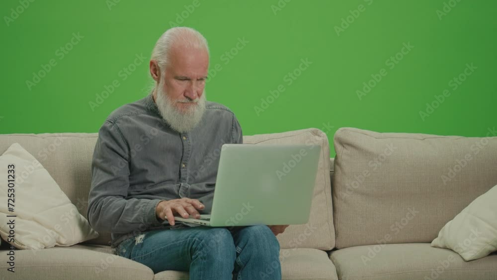Green Screen. A Smiling Old Man Sitting on a Sofa and Works on a Laptop. A Smart Elderly Man, Freelancer, Browsing on the Internet Using a Laptop. Virtual Communication for the Elderly. Green Screen. A Smiling Old Man Sitting on a Sofa and Works on a Laptop. A Smart Elderly Man, Freelancer, Browsing on the Internet Using a Laptop. Virtual Communication for the Elderly.