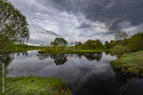 The river reflects the dramatic sky  a spring motif with the river and green bushes and grass.