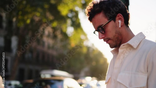 Pensive man in sunglasses and wireless earbuds listen audiobook while walk outdoors in slow motion. Confident male enjoy music from online playlist in earphones. Warm sunlight at summer evening

