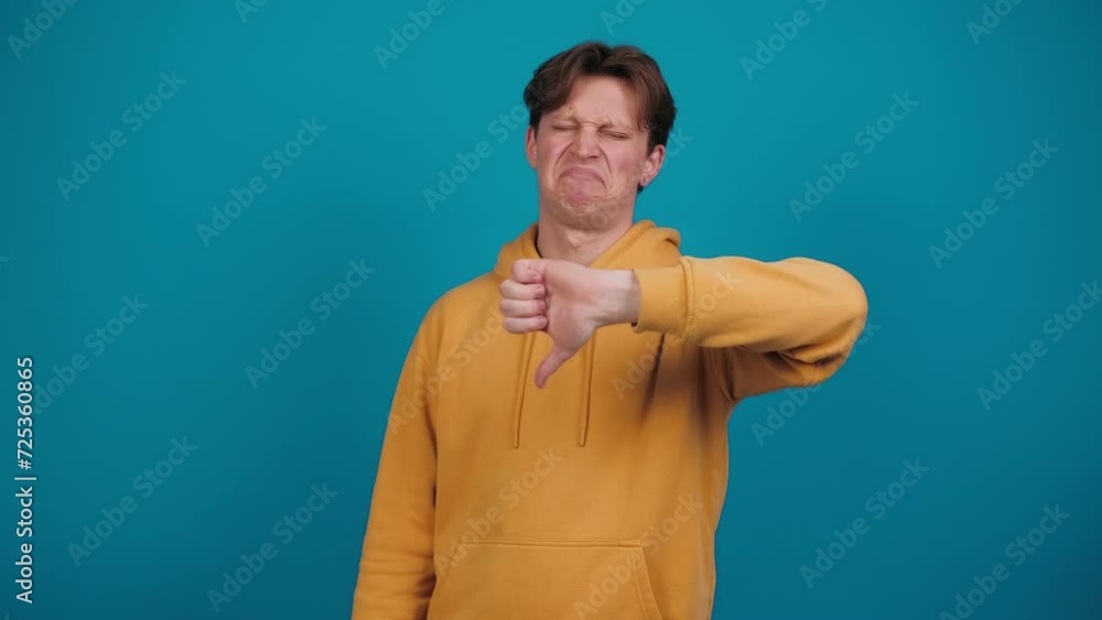 Dislike. Young man showing thumbs down sign gesture, expressing discontent, disapproval, dissatisfied negative feedback. Guy indoors isolated on blue background. Lifestyle. Emotions. People Dislike. Young man showing thumbs down sign gesture, expressing discontent, disapproval, dissatisfied negative feedback. Guy indoors isolated on blue background. Lifestyle. Emotions. People