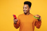 Young man wear orange sweatshirt casual clothes hold use mobile cell phone eat burger check calories isolated on plain yellow background. Proper nutrition healthy fast food unhealthy choice concept.