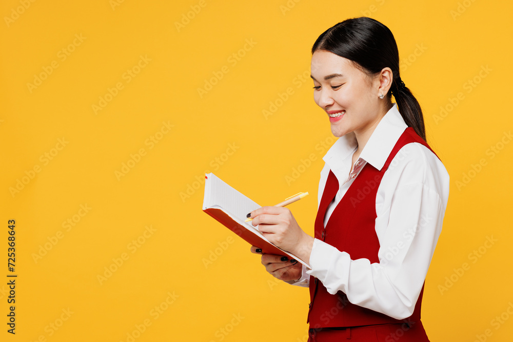 Side view young corporate lawyer employee business woman of Asian ethnicity wear formal red vest shirt work at office hold in hand diary notebook isolated on plain yellow background. Career concept. Side view young corporate lawyer employee business woman of Asian ethnicity wear formal red vest shirt work at office hold in hand diary notebook isolated on plain yellow background. Career concept.