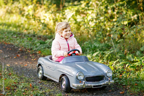 Little preschool girl driving big vintage toy car. Happy child having fun with playing outdoors. Active preschooler child enjoying warm autumn day in forest. Smiling stunning kid playing